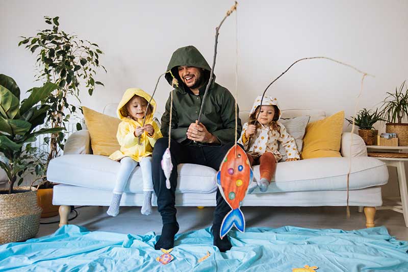 dad and 2 kids in rain jackets fake fishing indoors from couch.
